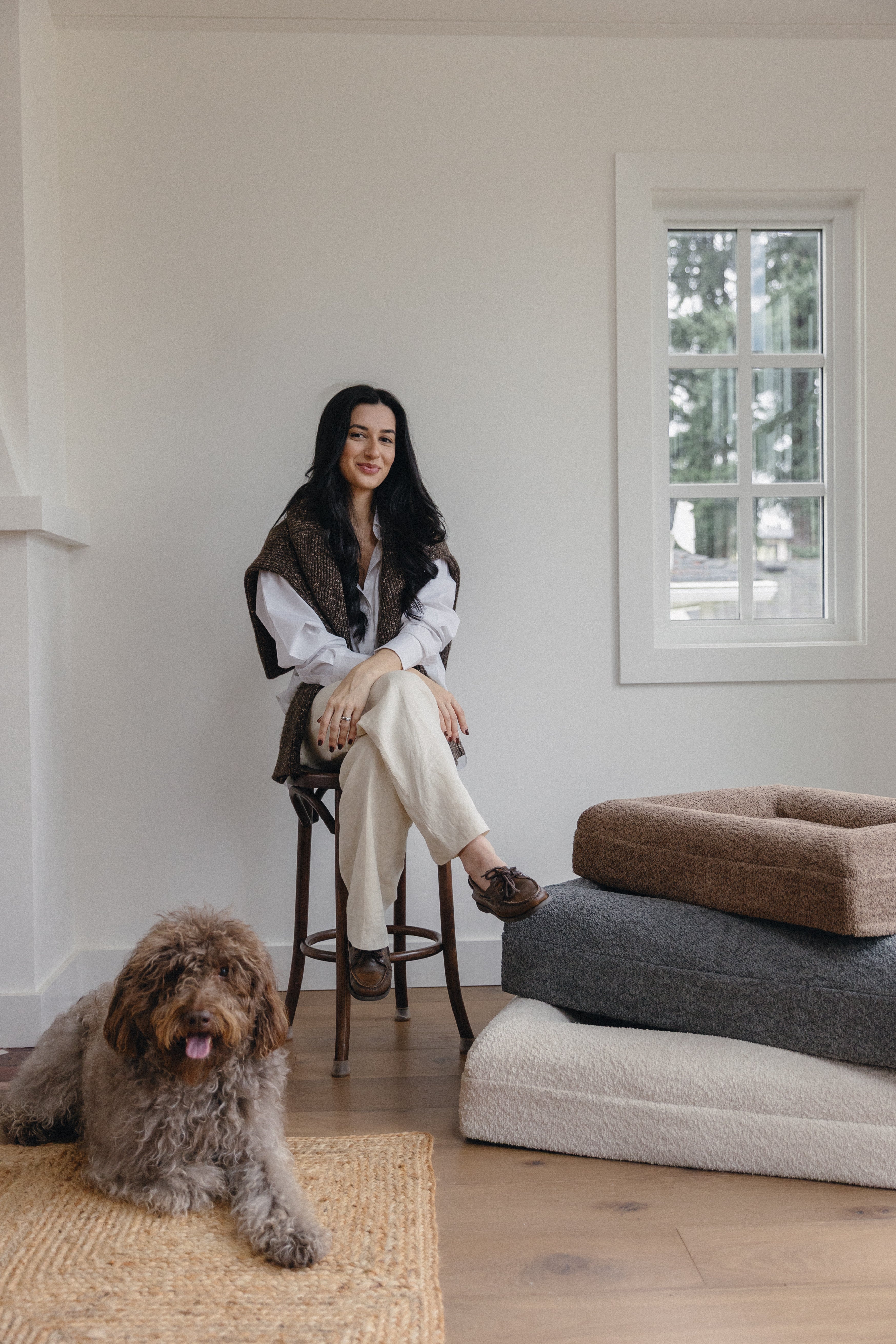 Woman sitting on a stool with a dog in a room with a window and furniture.